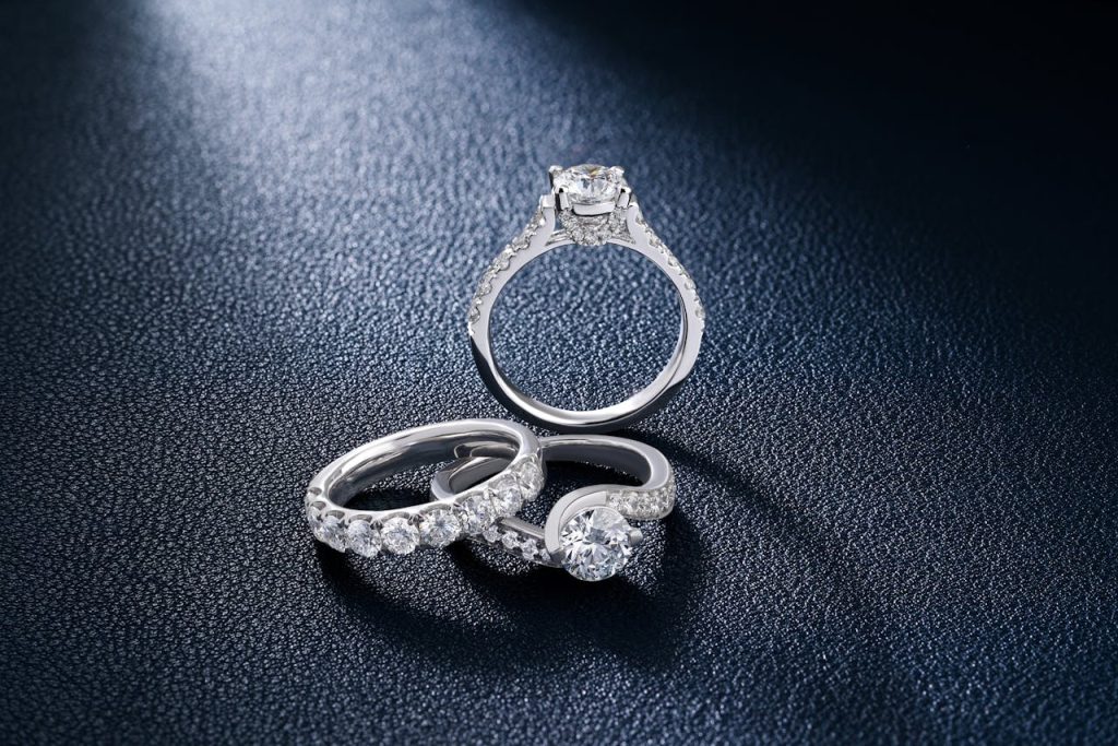 A collection of three exquisite diamond rings from One & Only displayed on a textured dark navy surface under a focused spotlight. The set includes a classic round-cut solitaire engagement ring with a pavé-set band, a full eternity wedding band featuring a continuous row of round diamonds, and a modern bypass-style diamond ring. The high-quality diamonds exhibit exceptional brilliance and fire, reflecting the brand's commitment to one-of-a-kind jewelry.